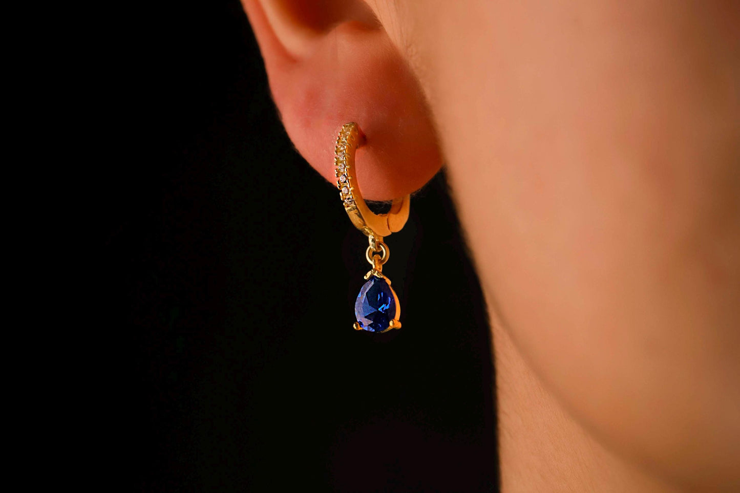 Gold earring with a blue gemstone worn on an ear against a black background