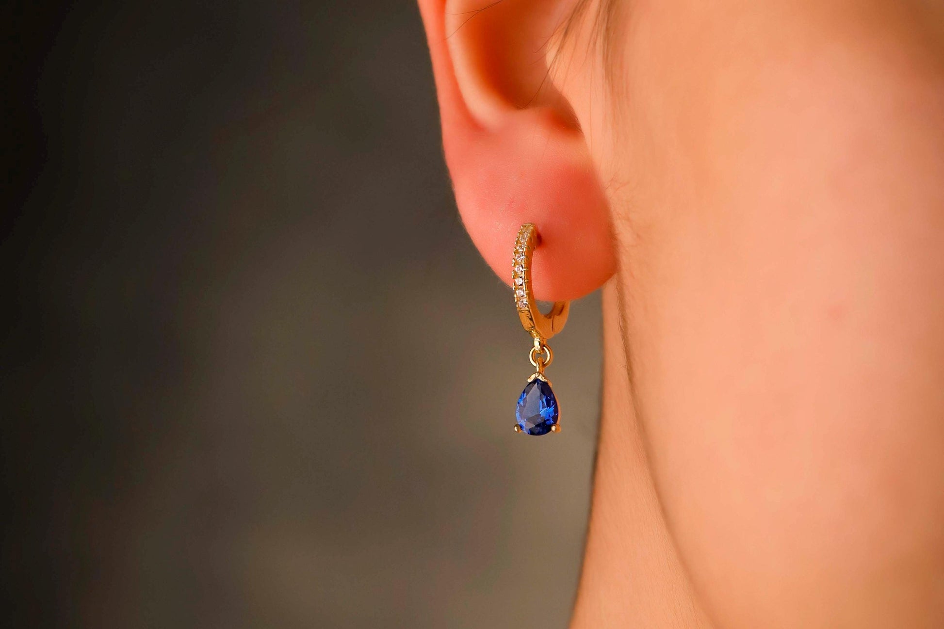 Gold earring with a blue gemstone worn by a person against a blurred background