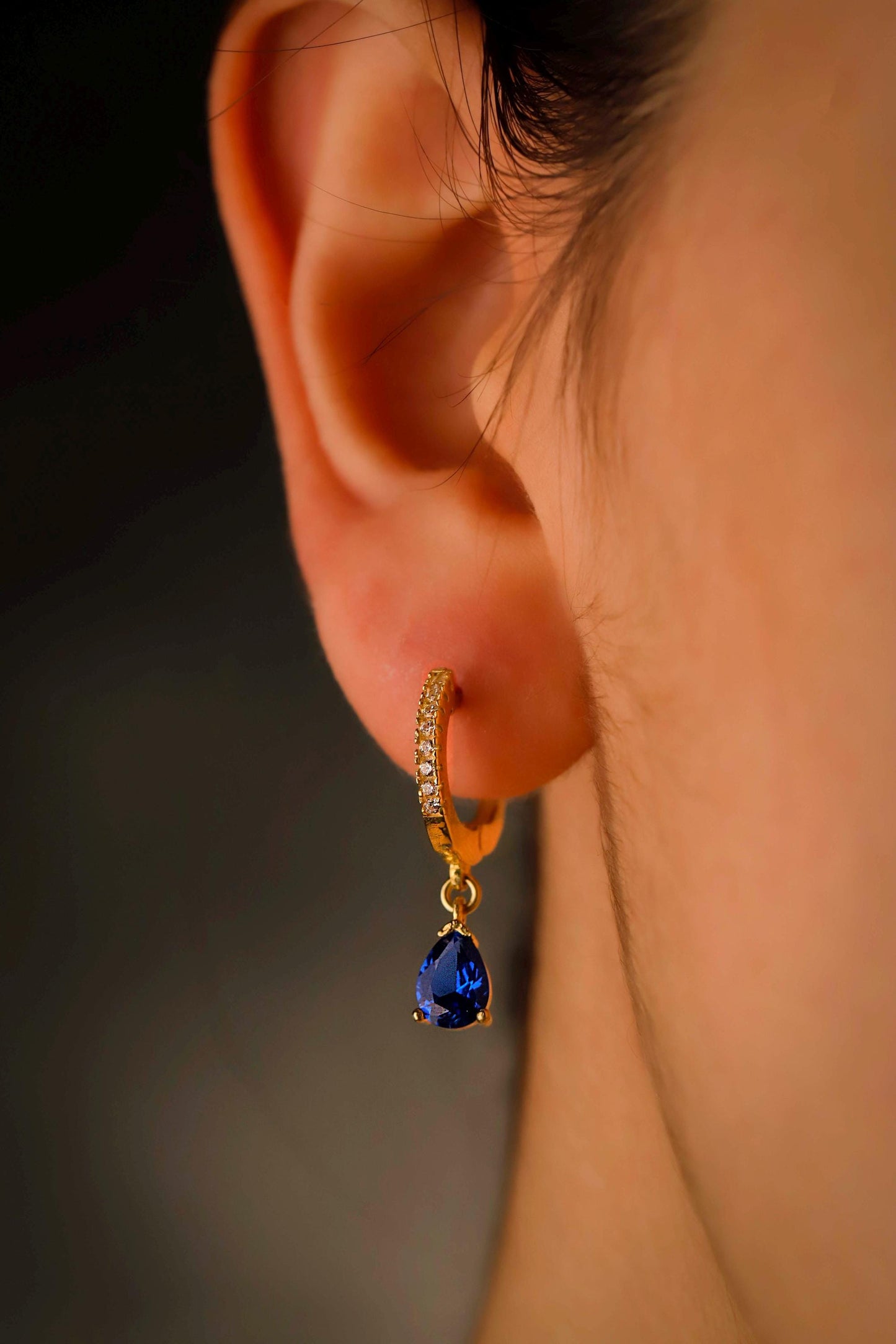 Close-up of an ear wearing a gold earring with a blue gemstone against a blurred background