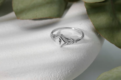 A silver initial ring with a small flower design, placed on a white surface with green leaves in the background.