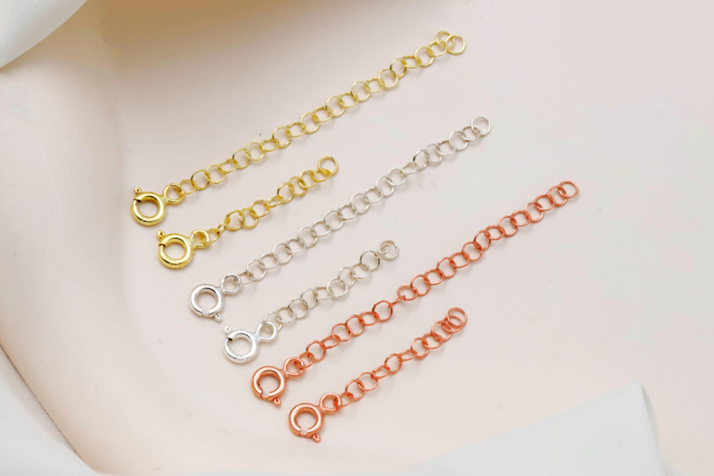 Three gold, silver, and rose gold chain necklaces on a white surface with greenery.