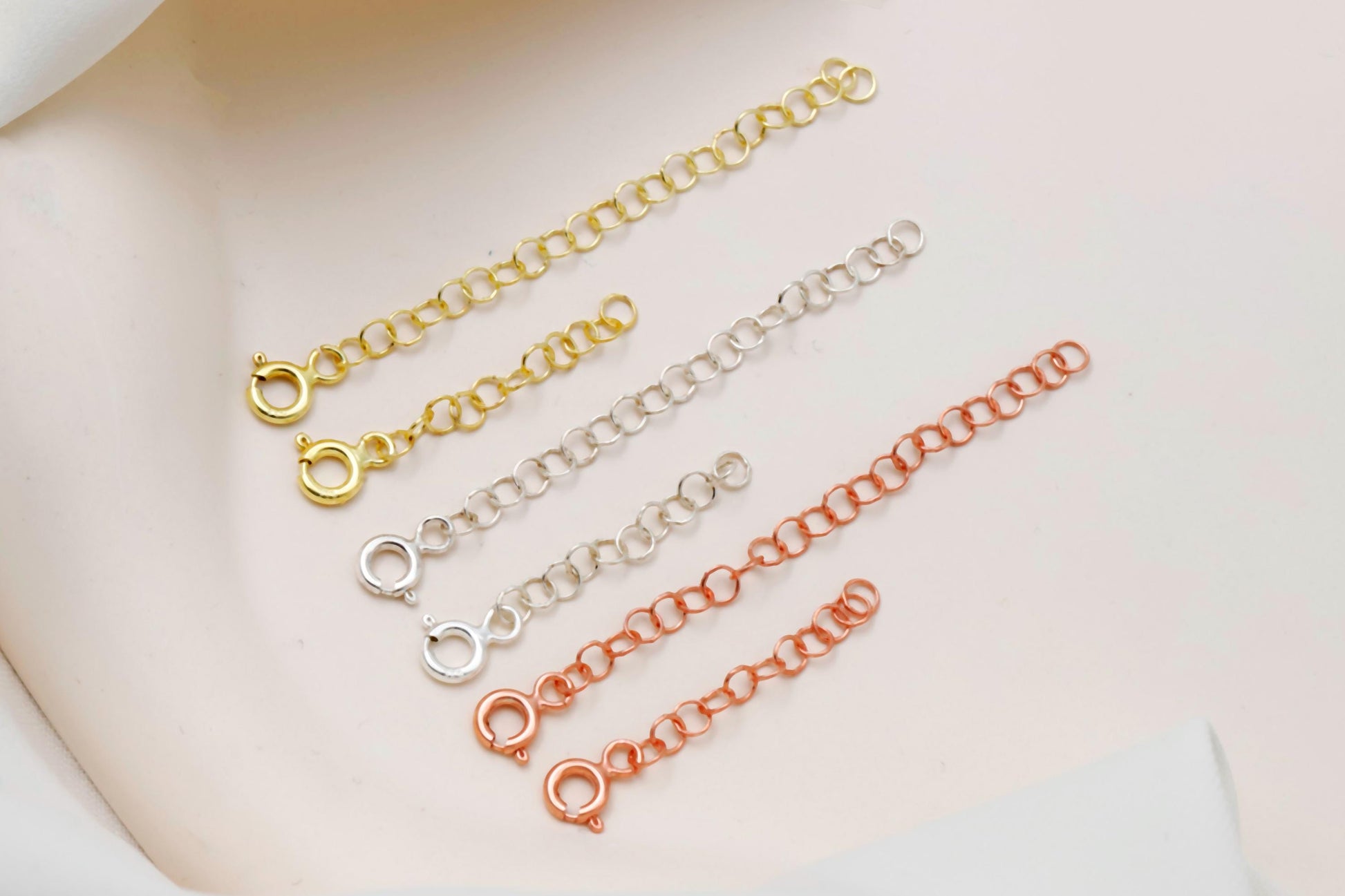 Three gold, silver, and rose gold chain necklaces on a white surface with greenery.