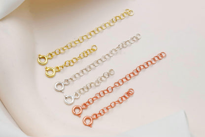 Three gold, silver, and rose gold chain necklaces on a white surface with greenery.