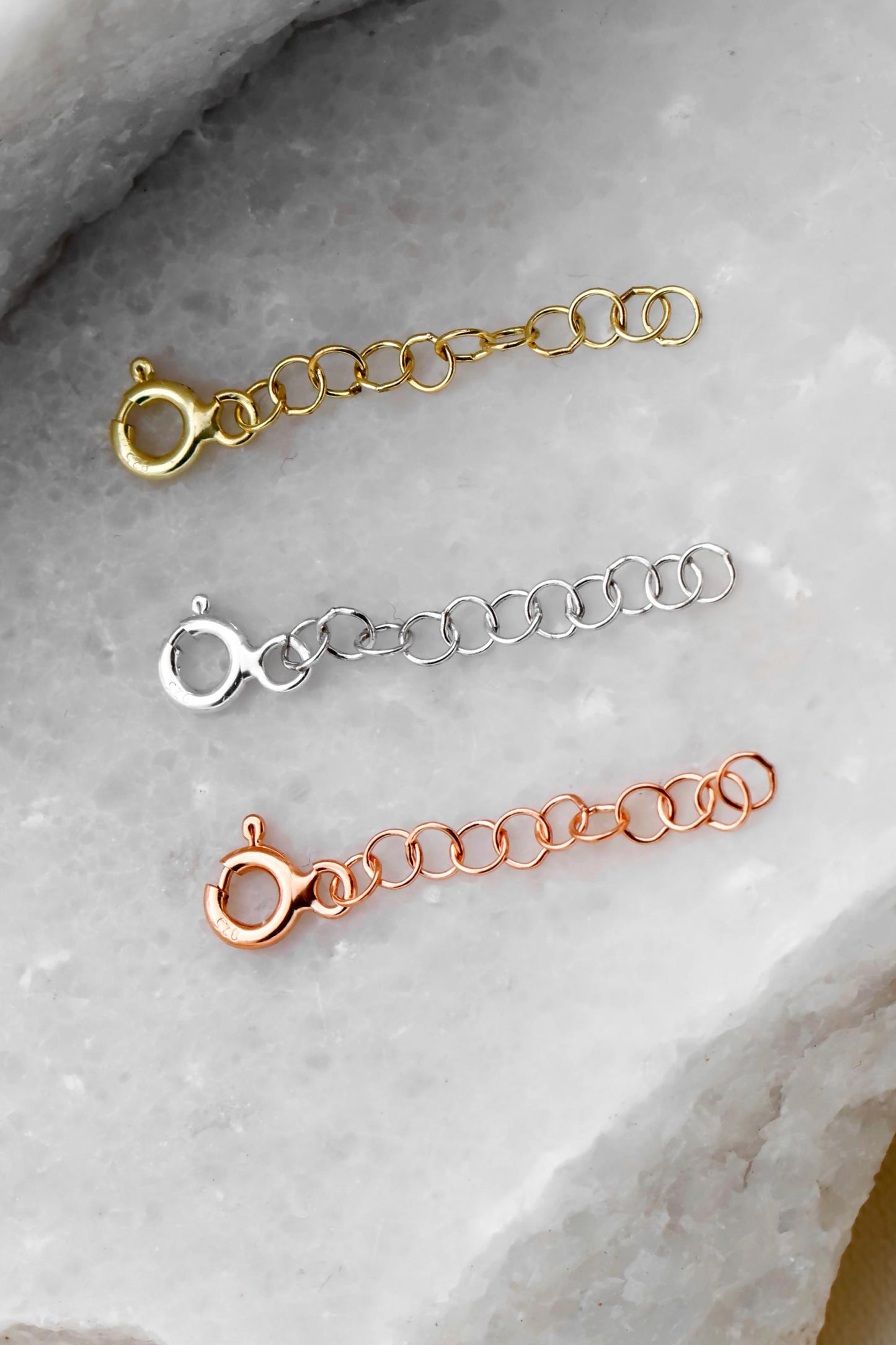 Three adjustable extender chains in gold, silver, and rose gold colors laid out on a marble surface with a decorative flower in the background.