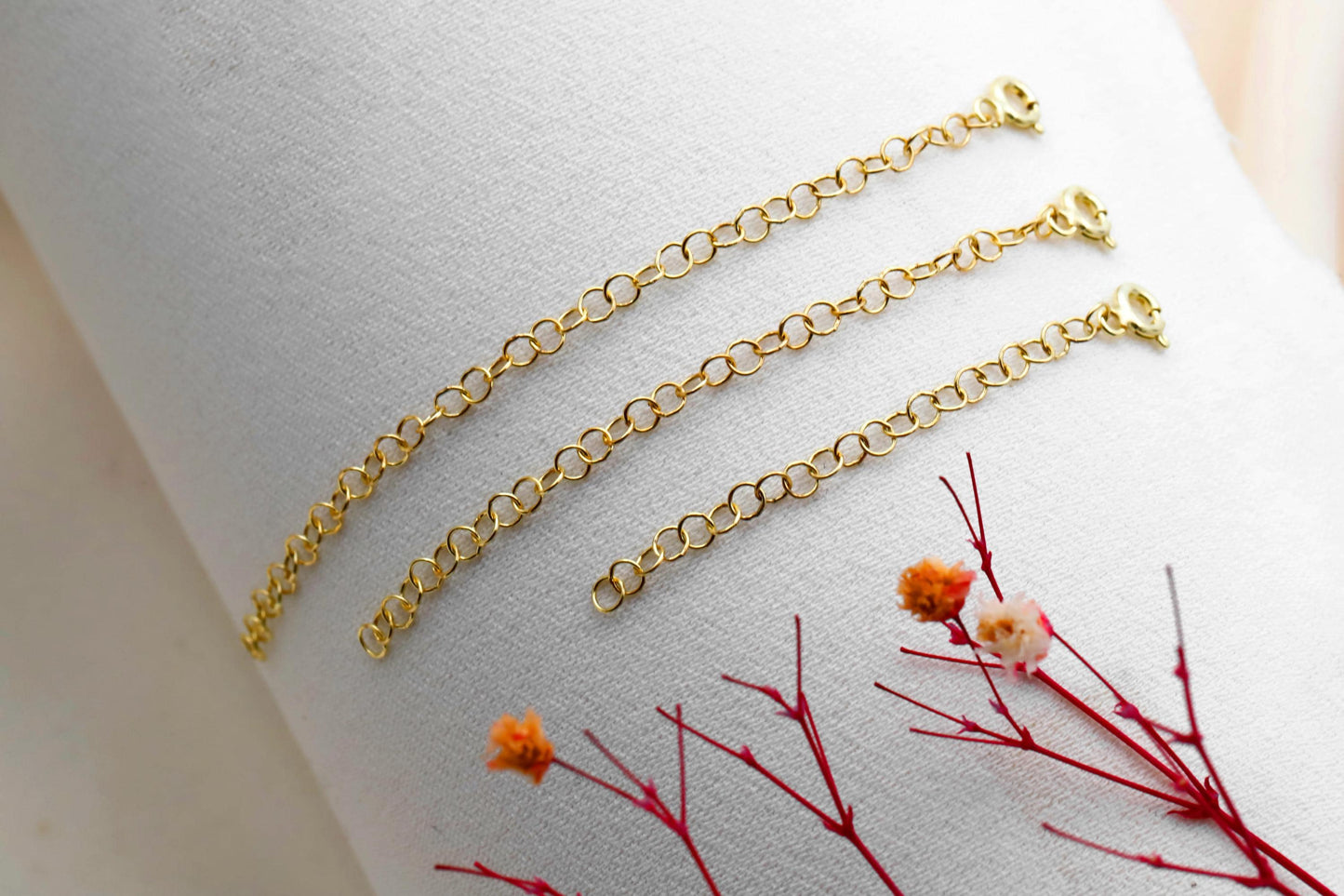 Three adjustable extender chains in gold, silver, and rose gold colors laid out on a marble surface with a decorative flower in the background.