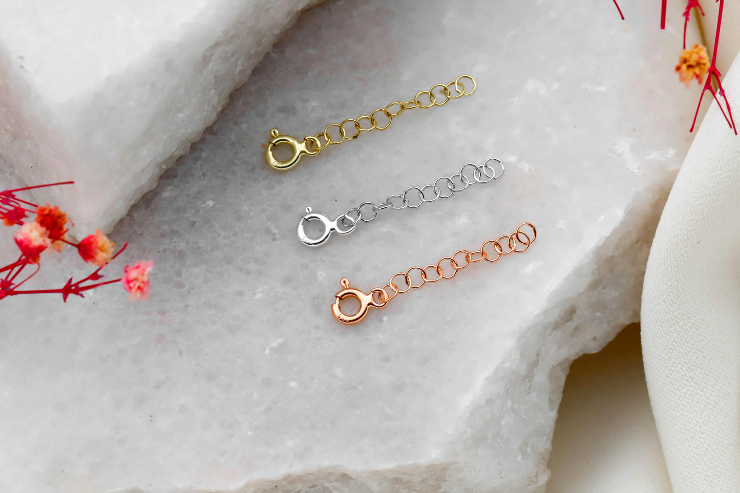 Three adjustable extender chains in gold, silver, and rose gold colors laid out on a marble surface with a decorative flower in the background.