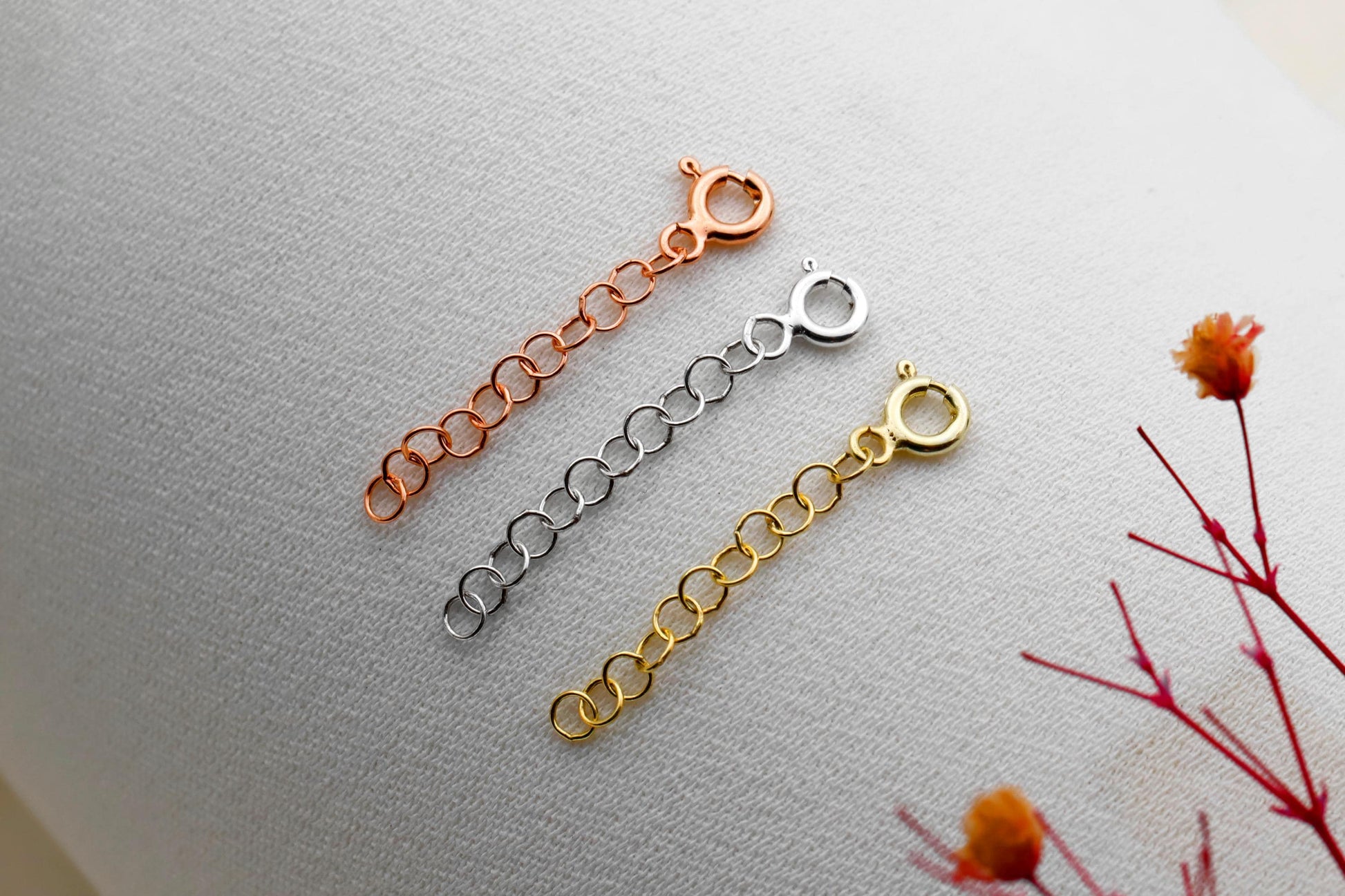 Three adjustable extender chains in gold, silver, and rose gold colors laid out on a marble surface with a decorative flower in the background.