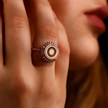 A close-up image of a person's hand showcasing a university ring with a round centerpiece diamond, surrounded by smaller diamonds, and inscriptions on the band.