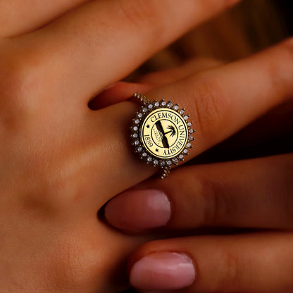 A close-up image of a person's hand showcasing a university ring with a round centerpiece diamond, surrounded by smaller diamonds, and inscriptions on the band.