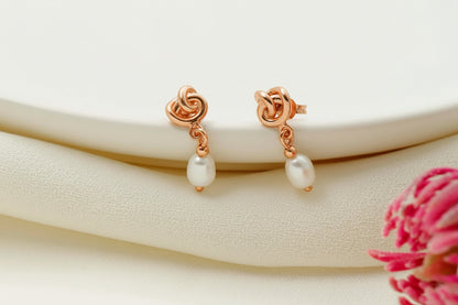 Rose gold earrings with pearl drops on a white surface with a pink flower.