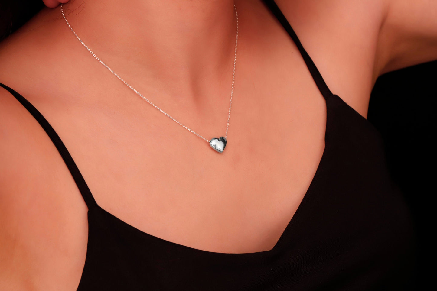 
A silver, heart-shaped pendant hangs from a thin silver chain on a person's neck. A product by SilverGoldandRose.com.
