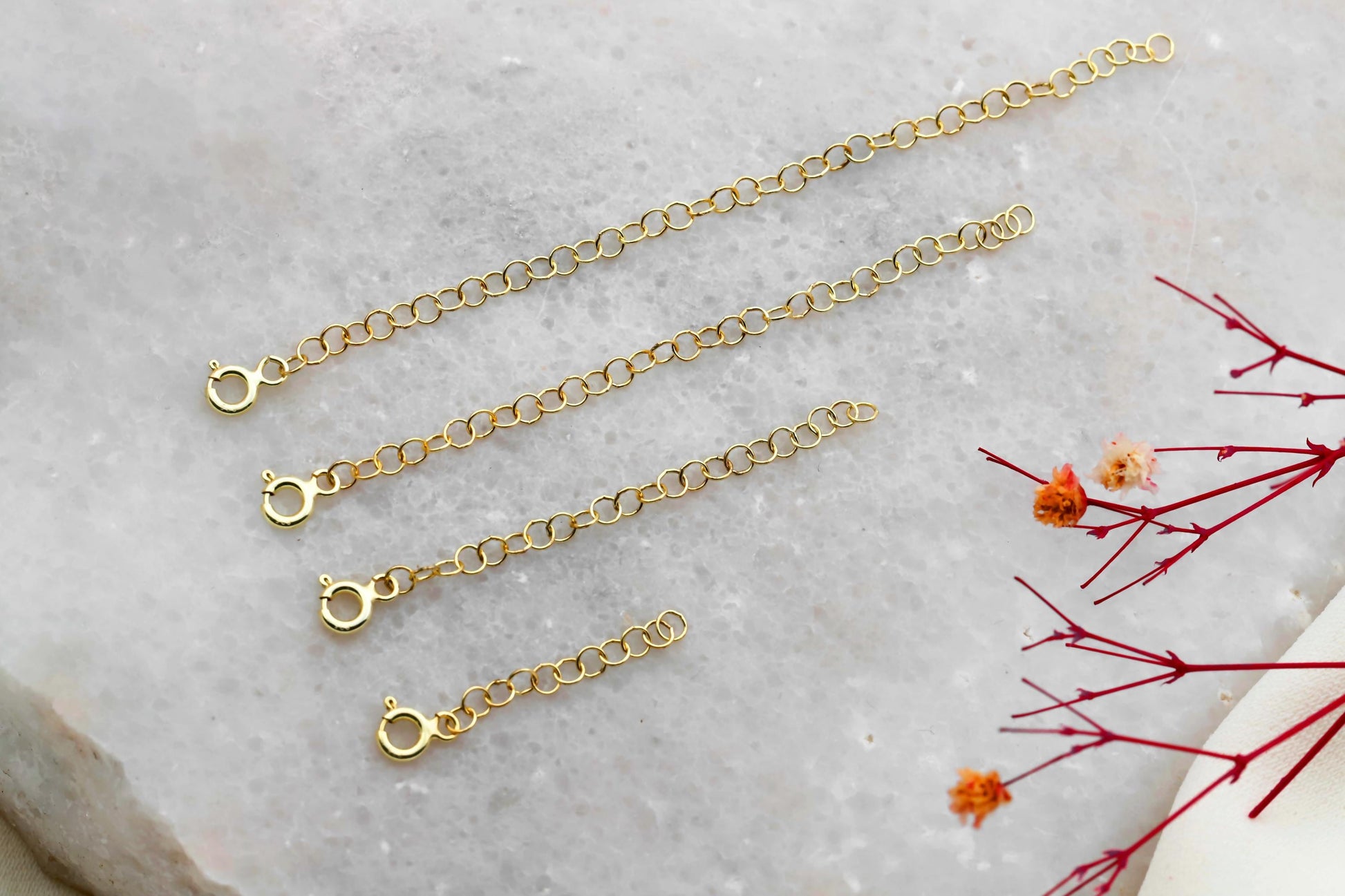 Three adjustable extender chains in gold, silver, and rose gold colors laid out on a marble surface with a decorative flower in the background.