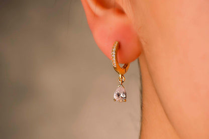 Gold hoop earring with a teardrop-shaped gemstone worn on an ear against a neutral background