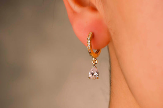 Gold hoop earring with a teardrop-shaped gemstone worn on an ear against a neutral background