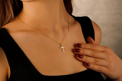 Gold necklace with a cross pendant worn by a person, with a hand touching the shoulder.