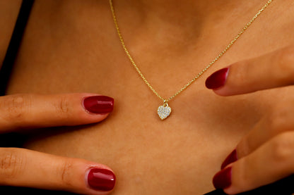 A close-up image of a woman wearing a 14K gold pave diamond heart necklace with a silver chain, showcased on a black dress.