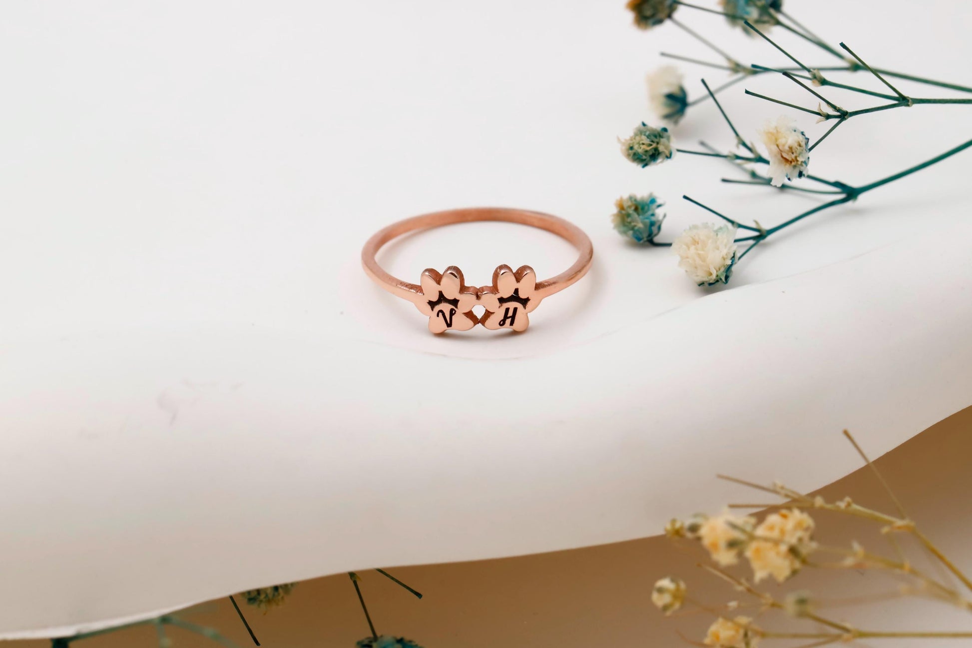Rose gold ring with paw prints on a white surface with dried flowers.