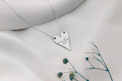 Heart-shaped silver necklace with engraved text on a white surface with dried flowers.