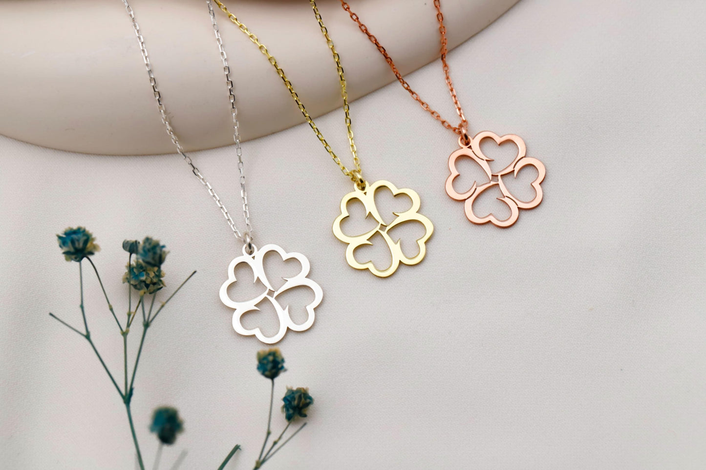 Four clover-shaped pendants on necklaces in silver, gold, and rose gold, with dried flowers on a light background.