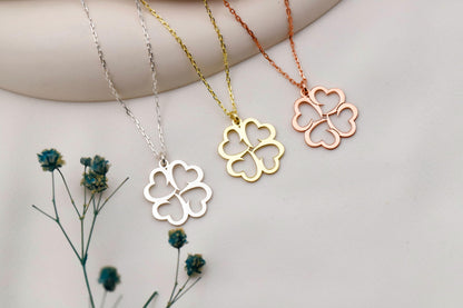 Four clover-shaped pendants on necklaces in silver, gold, and rose gold, with dried flowers on a light background.