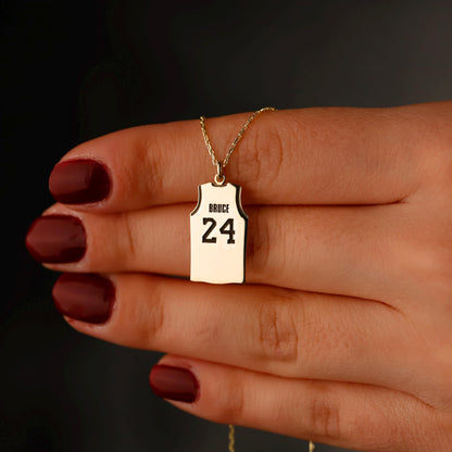 Gold necklace with a pendant shaped like a basketball jersey held by a hand against a dark background
