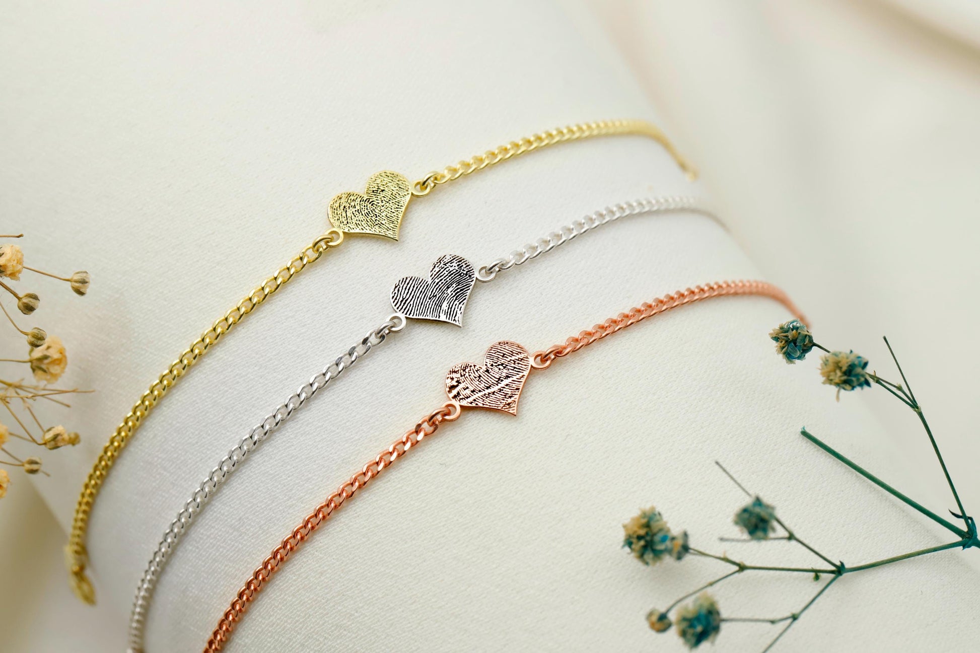 Three gold, silver, and rose gold bracelets on a white surface with dried flowers.