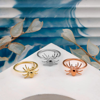 Three spider-shaped rings in gold, silver, and rose gold on a white stand with a blue and white striped background.