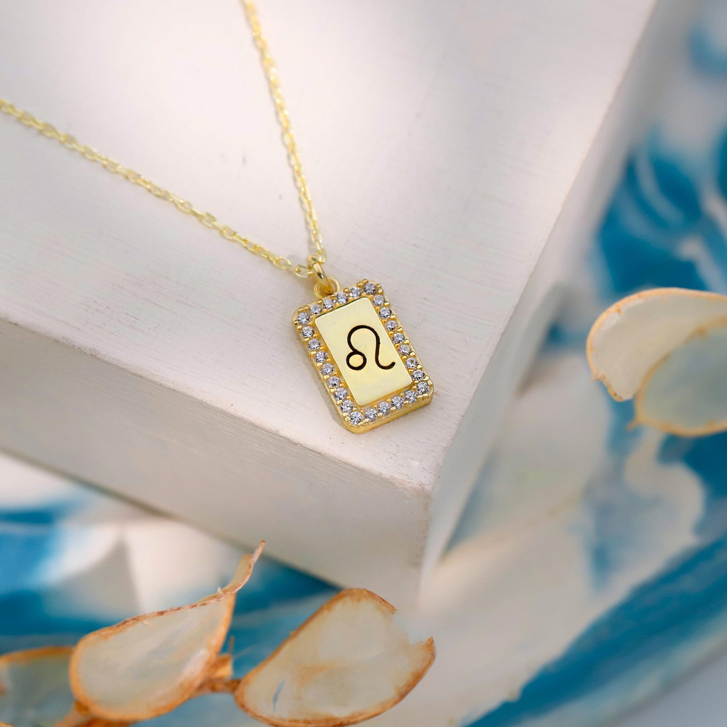 Gold necklace with a rectangular pendant featuring a symbol, on a white surface with dried leaves.