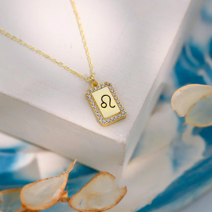 Gold necklace with a rectangular pendant featuring a symbol, on a white surface with dried leaves.