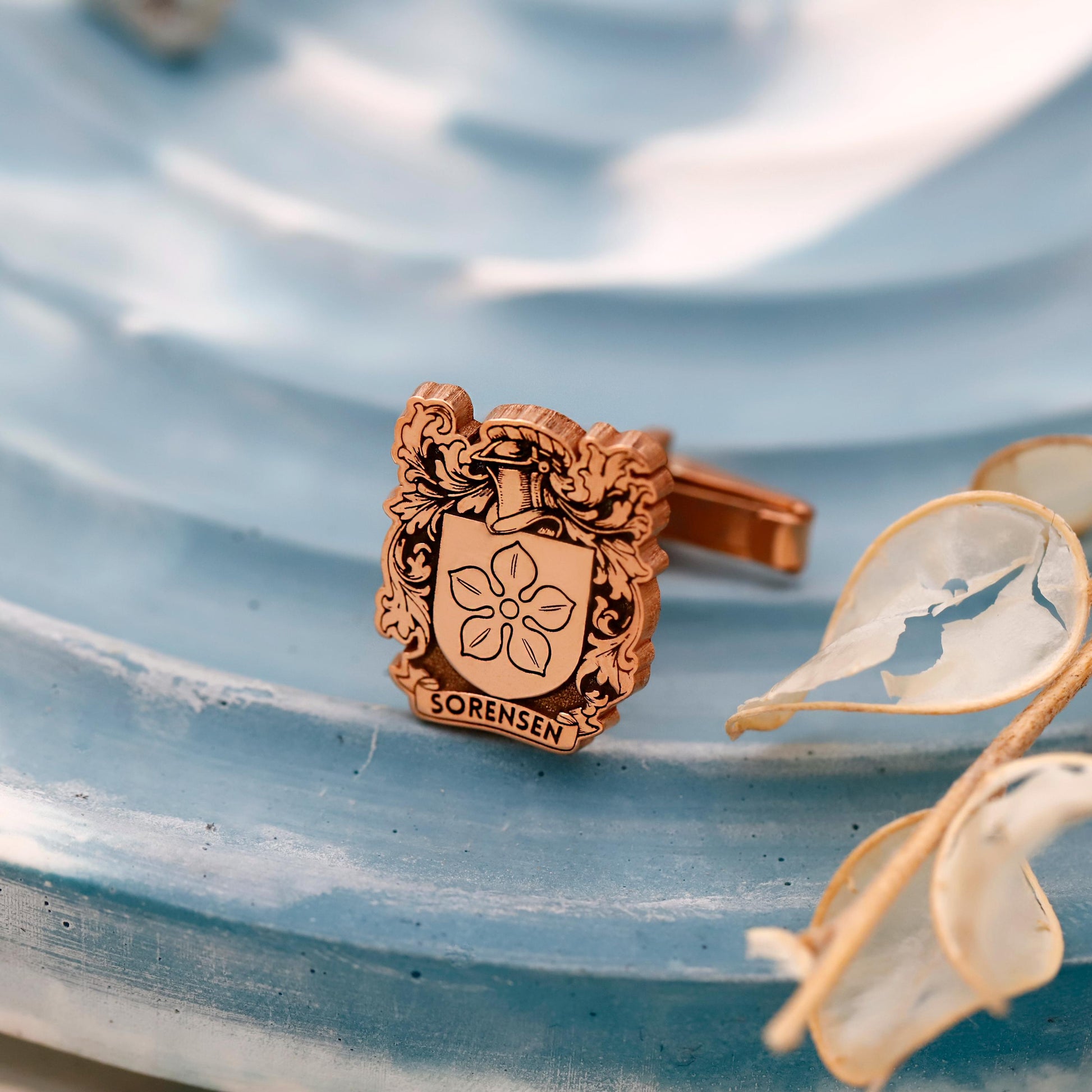 Cufflink with floral design and 'Sorensen' engraving on a textured blue surface with dried flowers.