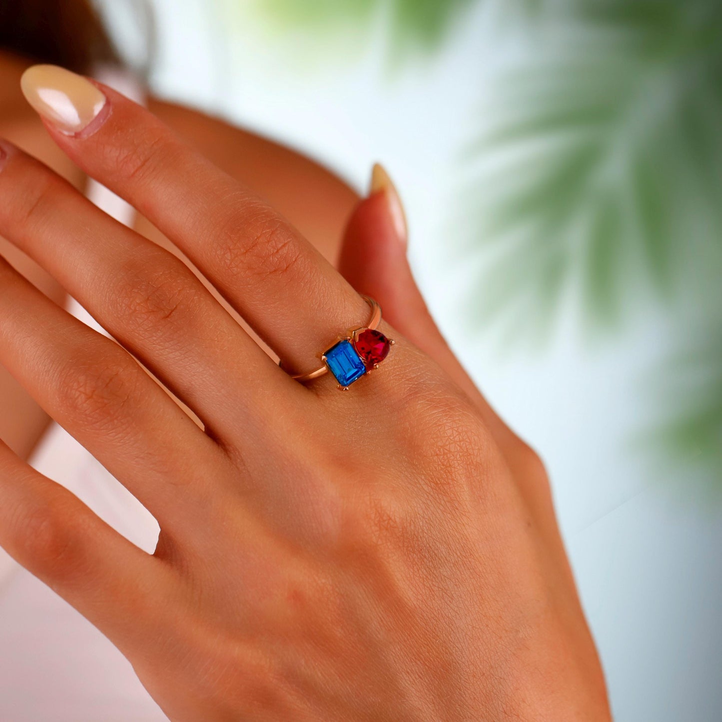 Close-up of a hand wearing a ring with red and blue gemstones against a blurred natural background