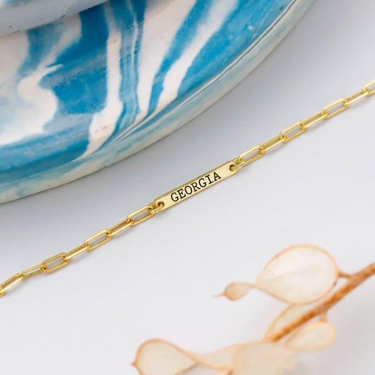 Gold bracelet with 'GEORGIA' engraving on a white surface with a blue and white patterned object in the background.