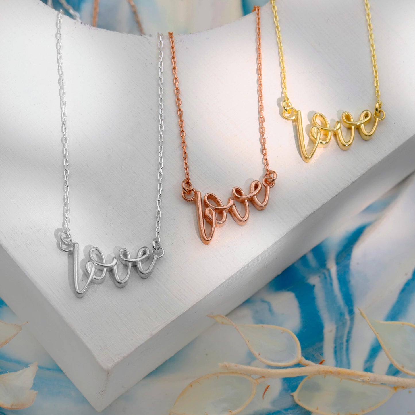 Three necklaces with 'love' inscription in silver, rose gold, and gold on a white stand with a blue floral background.