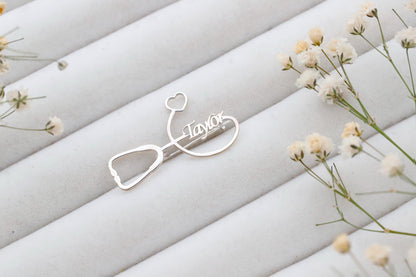 Silver stethoscope-shaped hair clip with 'Taylor' engraving on a light fabric background with small flowers.
