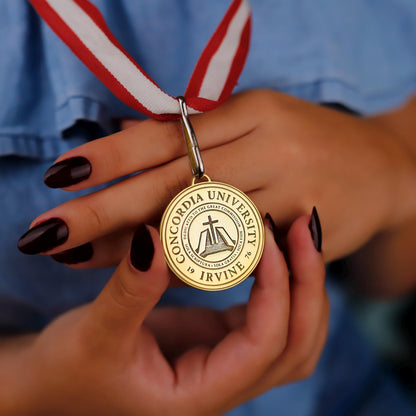 Gold medal with Concordia University Irvine logo held by a person wearing a blue shirt.