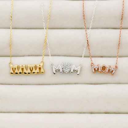 Three necklaces with 'MOM' text in gold, silver, and rose gold on a soft beige background.