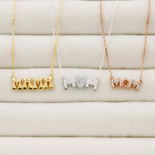 Three necklaces with 'MOM' text in gold, silver, and rose gold on a soft beige background.