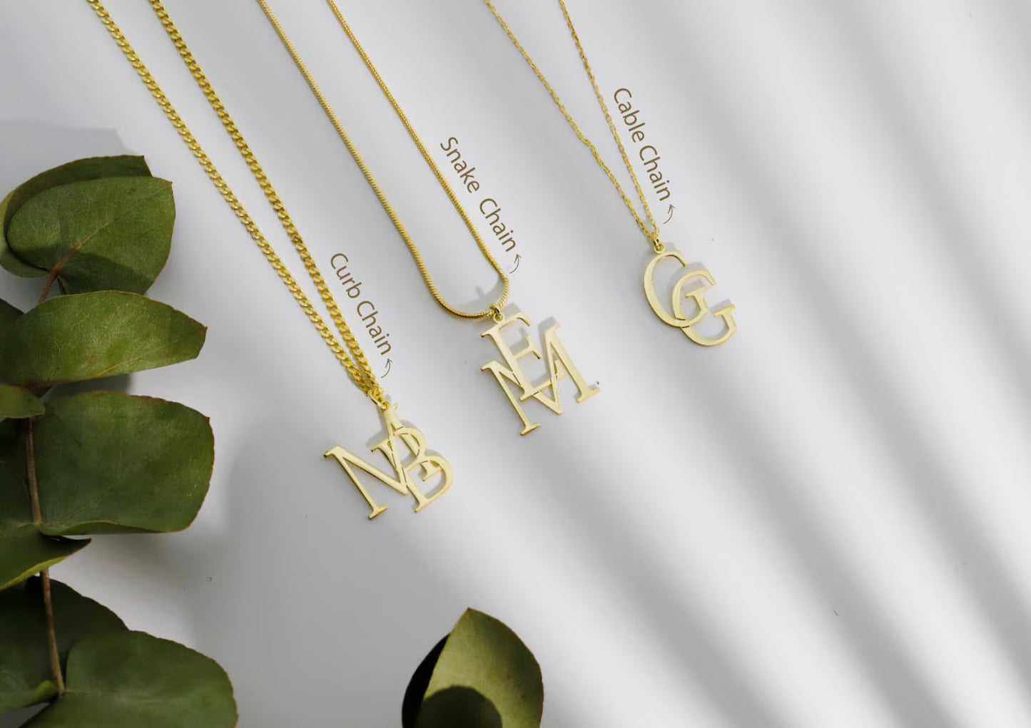 Gold necklaces with different chains and initials on a white background with green leaves.
