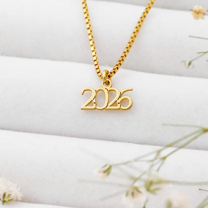 Gold necklace with '2026' pendant on a white surface with flowers.