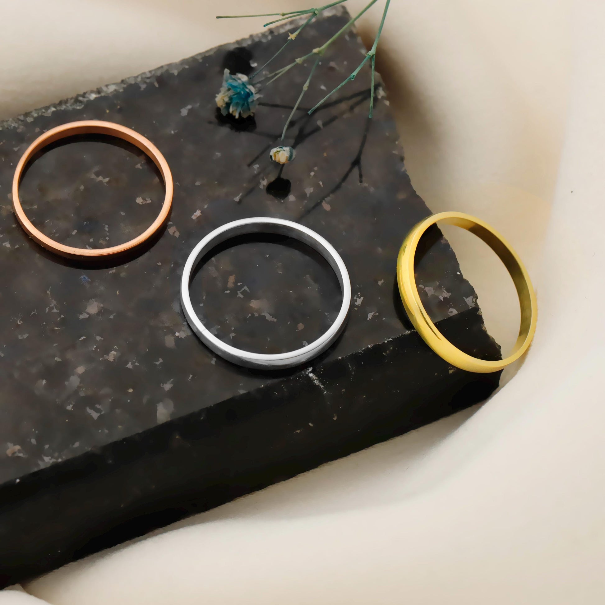 Three metallic rings (bronze, silver, gold) on a dark stone surface with a minimal background.