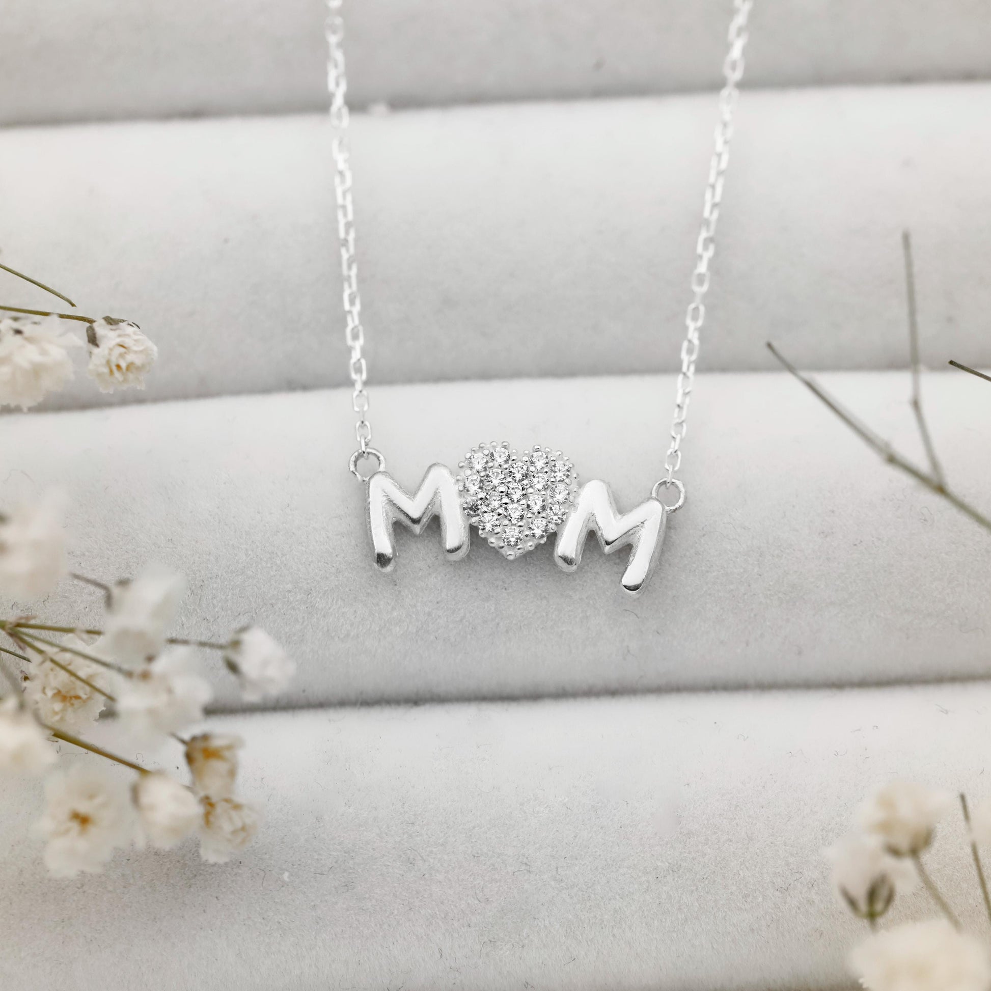 Silver necklace with 'MOM' and heart pendant on a light gray surface with white flowers.