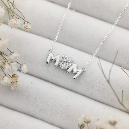 Silver necklace with 'MOM' pendant on a light gray surface with white flowers.
