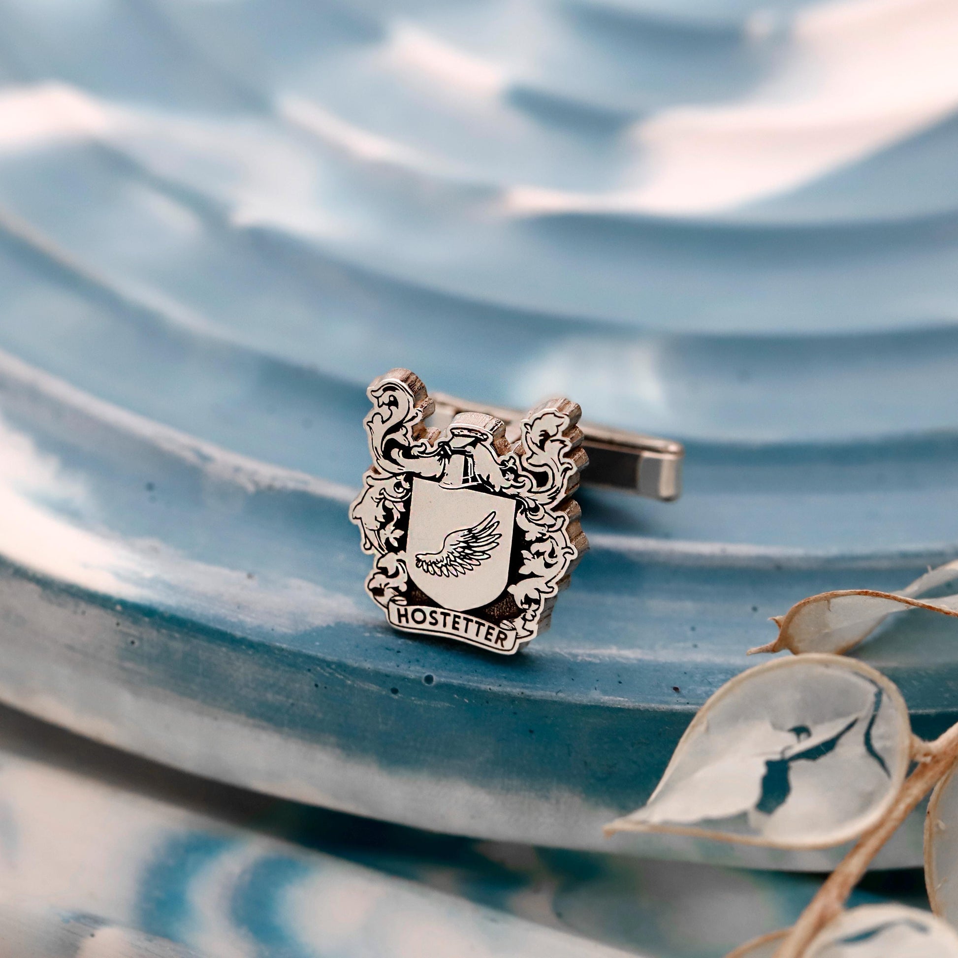 Silver crest-shaped cufflinks with 'Hostetter' on a blue textured background