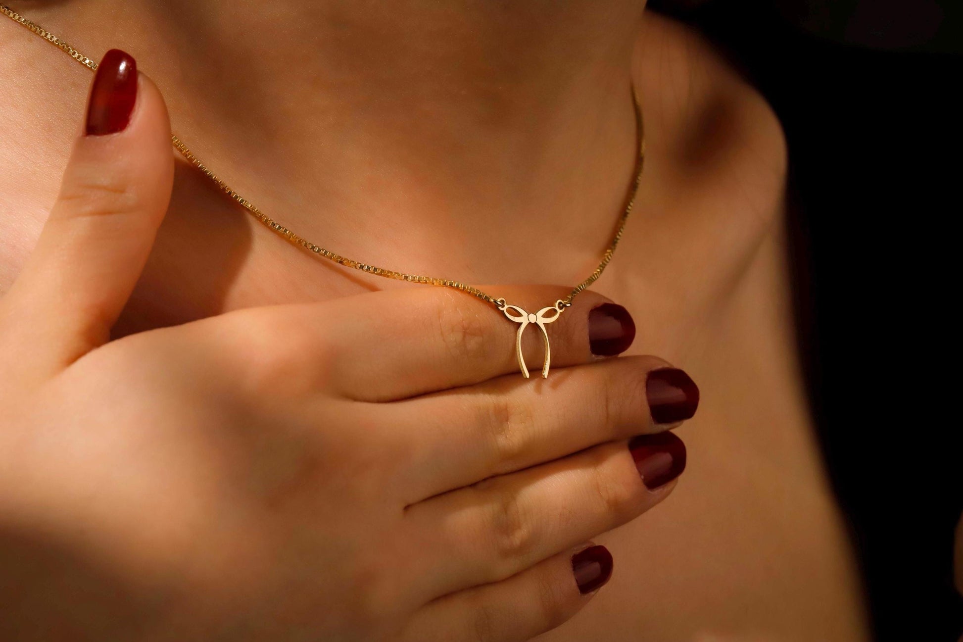 Gold necklace with a bow pendant held by hands with red nail polish.