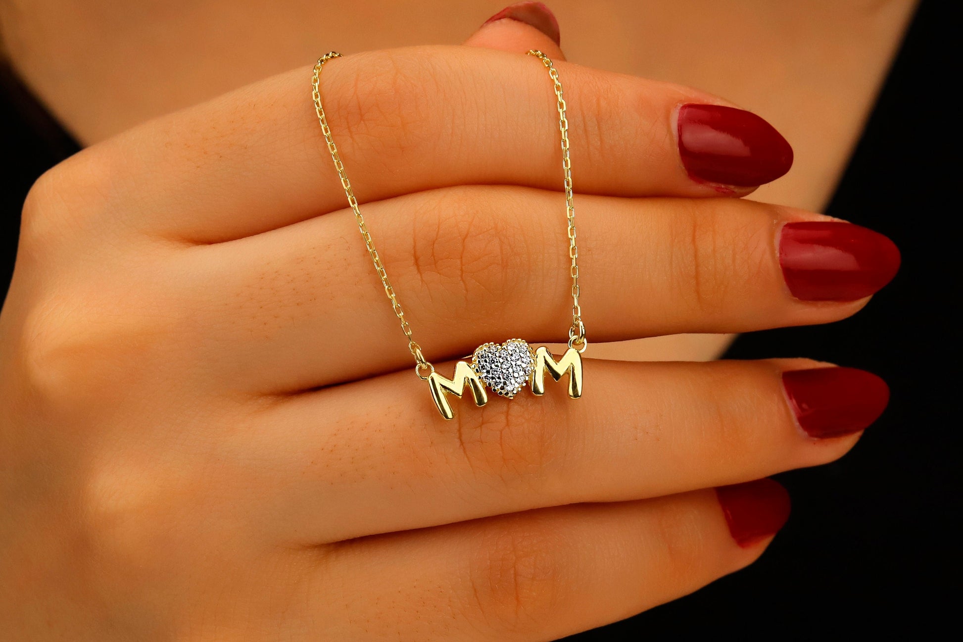 Gold necklace with a heart and 'MOM' pendant on a hand with red nail polish.