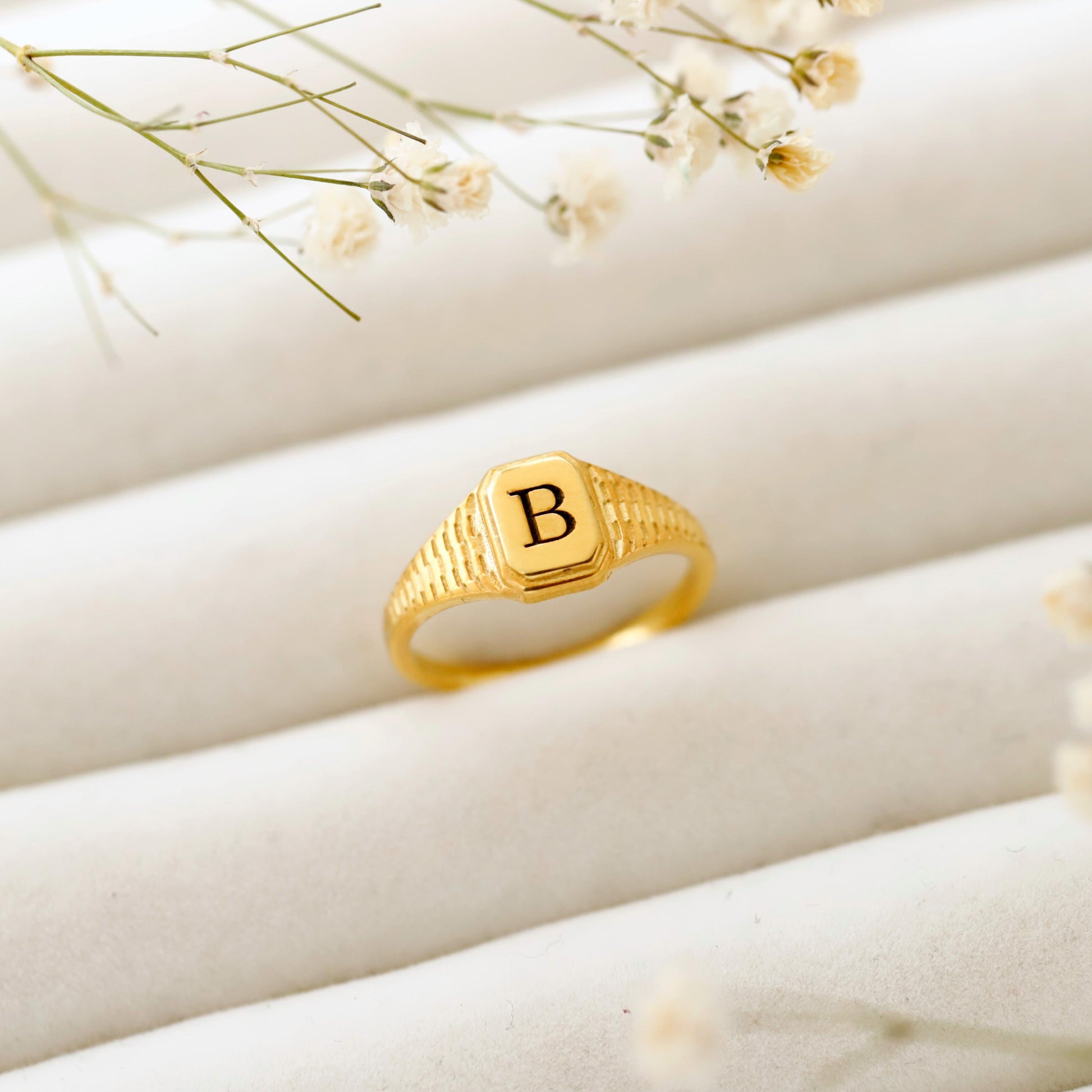 Gold ring with letter 'B' on a beige surface with delicate flowers