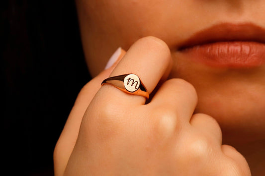 Close-up of a hand wearing a ring with a 'M' symbol, against a blurred background.