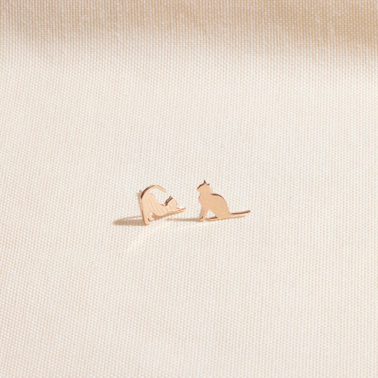 A gold cat stud earring in a playful stretching pose, shown held on a fingertip and being worn on an ear. A product by SilverGoldandRose.com.
