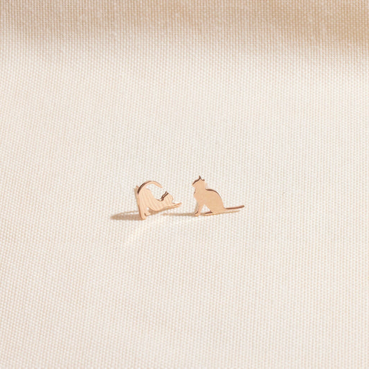 A gold cat stud earring in a playful stretching pose, shown held on a fingertip and being worn on an ear. A product by SilverGoldandRose.com.