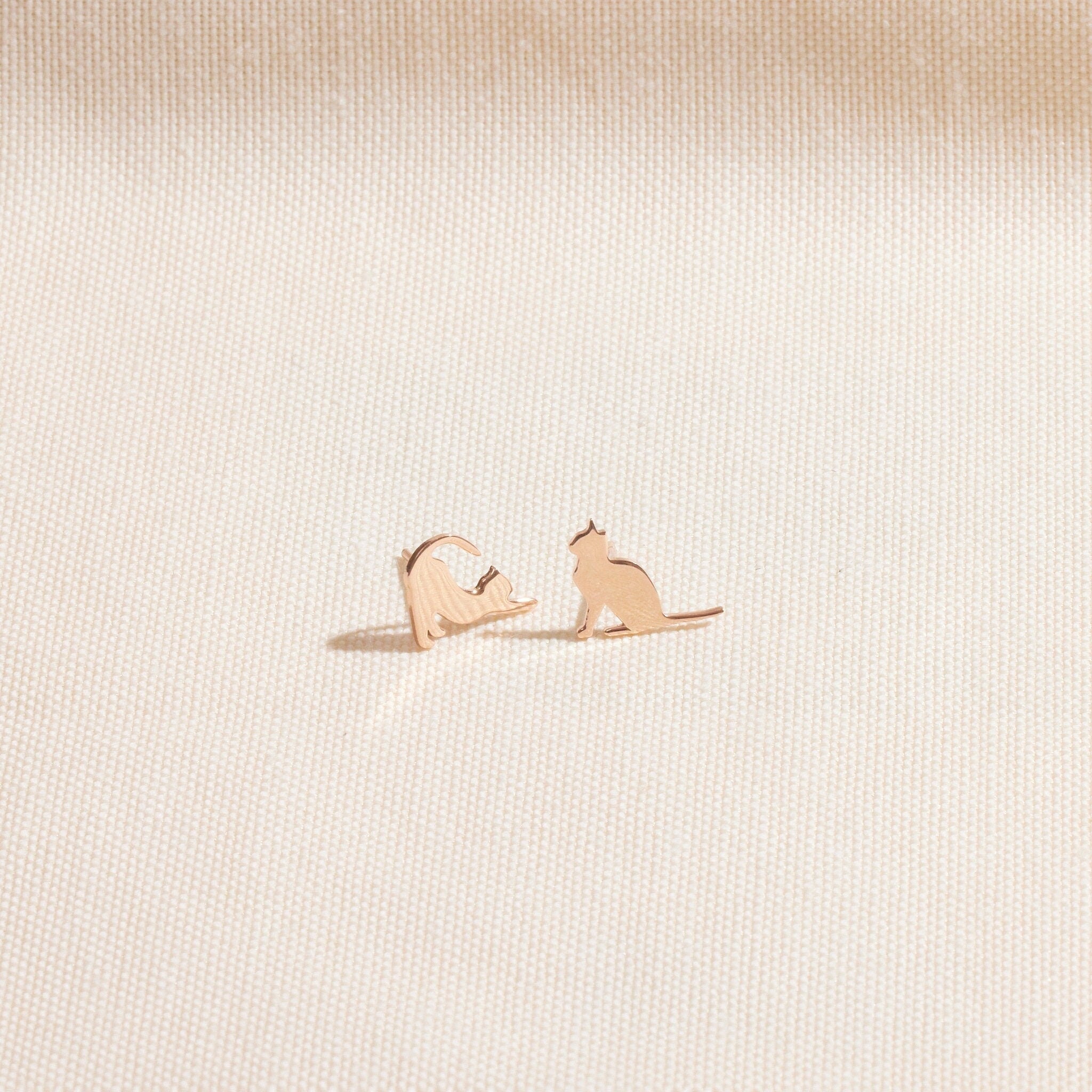 A gold cat stud earring in a playful stretching pose, shown held on a fingertip and being worn on an ear. A product by SilverGoldandRose.com.